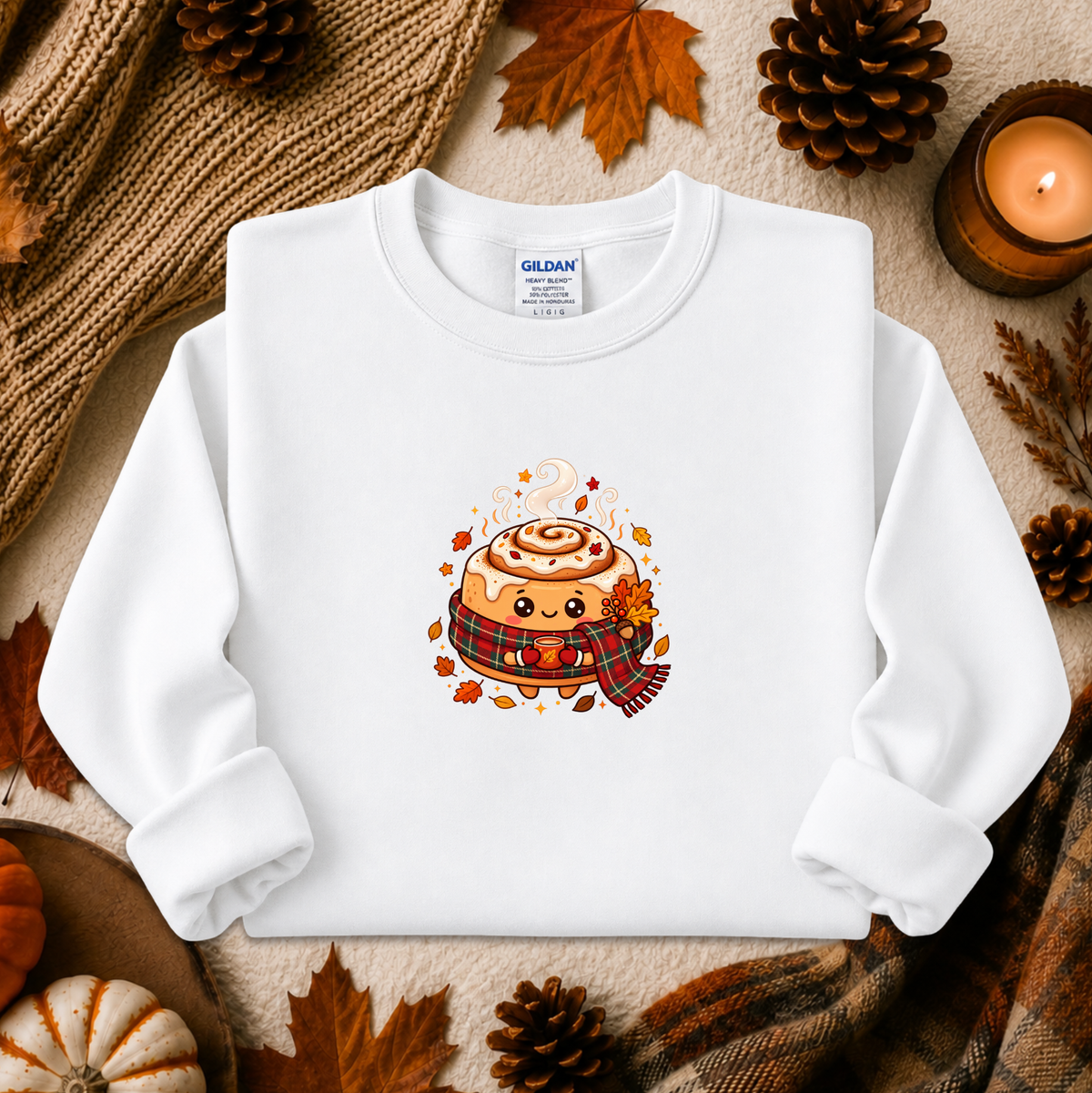 A white long-sleeve crewneck sweatshirt laid flat on a cozy autumn-themed background with pinecones, leaves, a candle, and a knitted scarf. The sweatshirt features a colorful cartoon graphic of a cinnamon roll character wearing a plaid scarf.