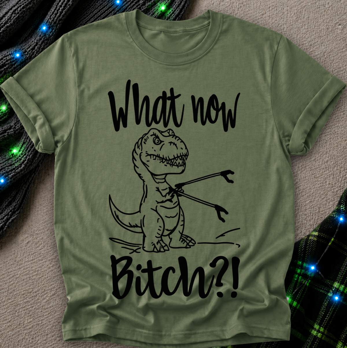 An olive green short-sleeve t-shirt with a black graphic of a cartoon T-Rex holding a fork and the text 'What now Bitch?!' printed on the front, laid flat on a textured beige surface with decorative blue string lights.