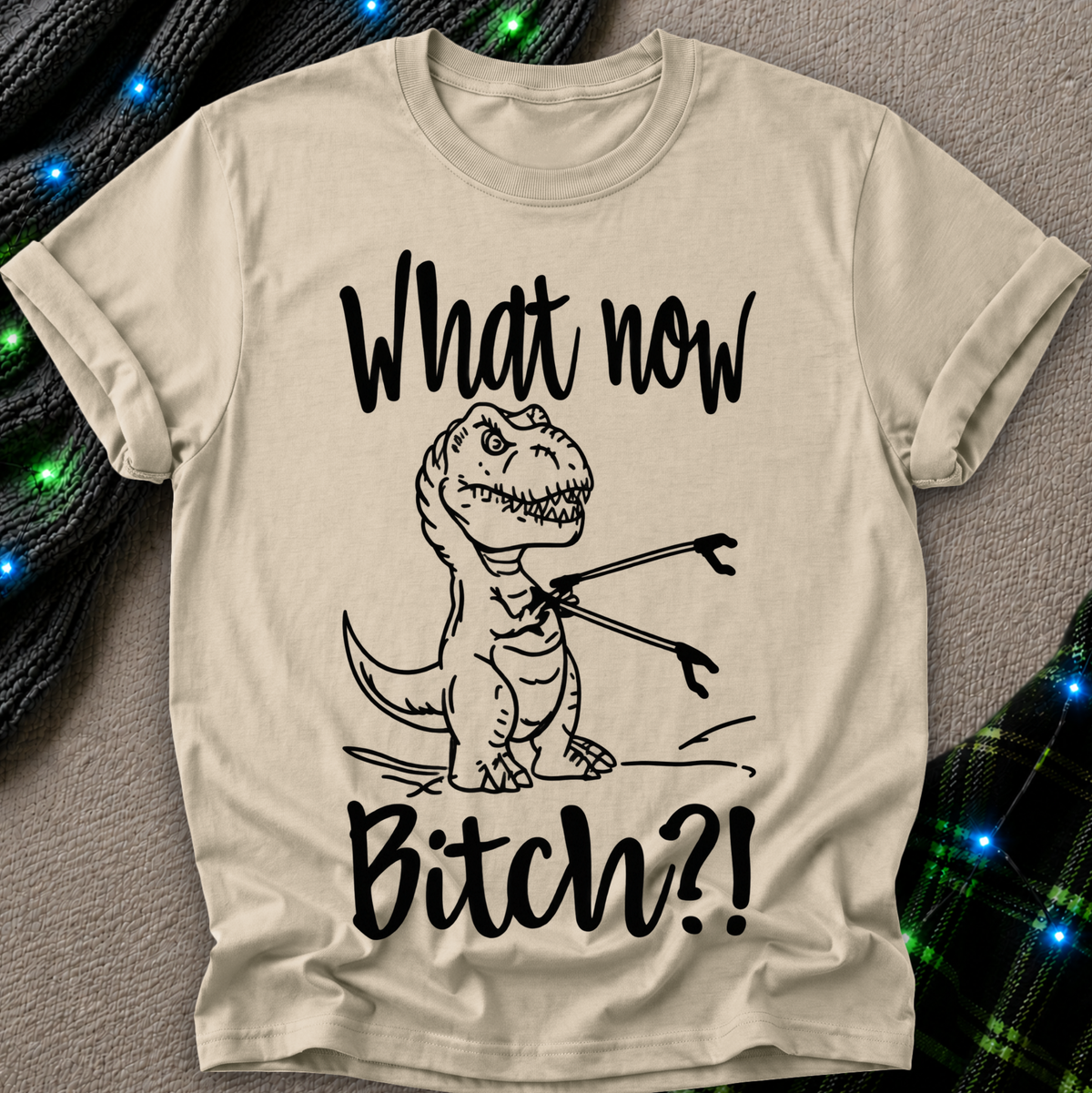 A beige graphic t-shirt featuring a black line art illustration of a T-Rex holding a fork, with the text 'What now Bitch?!' printed in black. The t-shirt is laid flat on a textured gray background with decorative green and black plaid fabric and blue string lights in the corners.