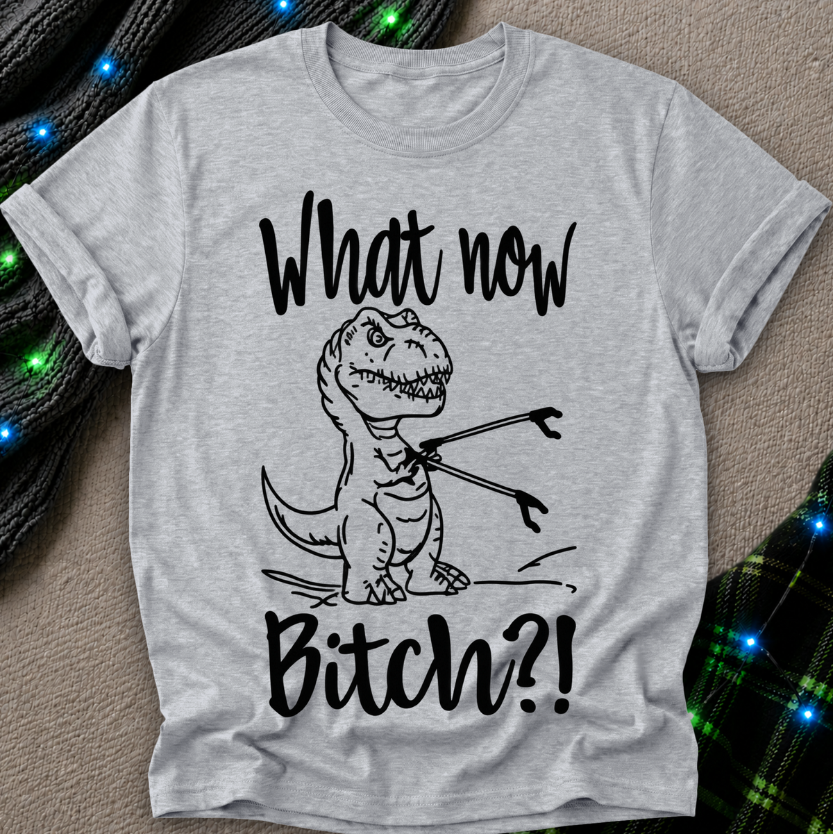 A light heather gray short-sleeve t-shirt with a black graphic of a cartoon T-rex holding a fork, with the text 'What now Bitch?!' printed around it. The shirt is laid flat on a textured beige surface with decorative green and black plaid fabric and blue string lights in the corners.