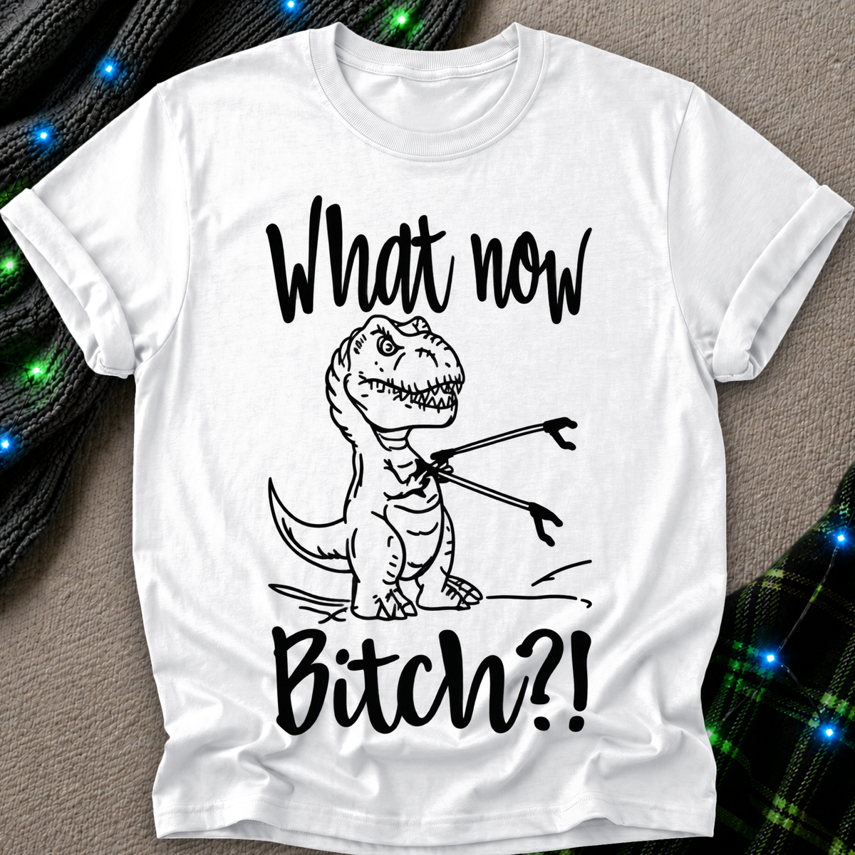 A white short-sleeve graphic T-shirt laid flat on a textured neutral background with decorative green and blue lights and plaid fabric at the corners. The shirt has a black line drawing of a cartoon T-rex holding a fork, with the text 'What now Bitch?!' in black script.