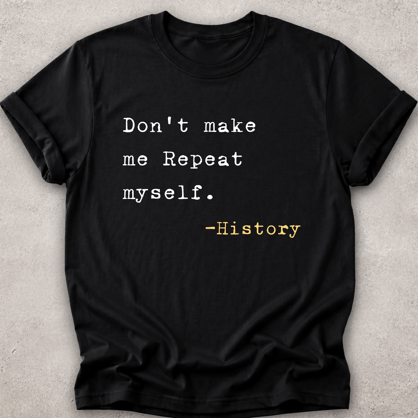 A black short-sleeve crewneck t-shirt laid flat on a textured light gray background. The shirt features a typewriter-style printed quote that reads 'Don't make me Repeat myself.