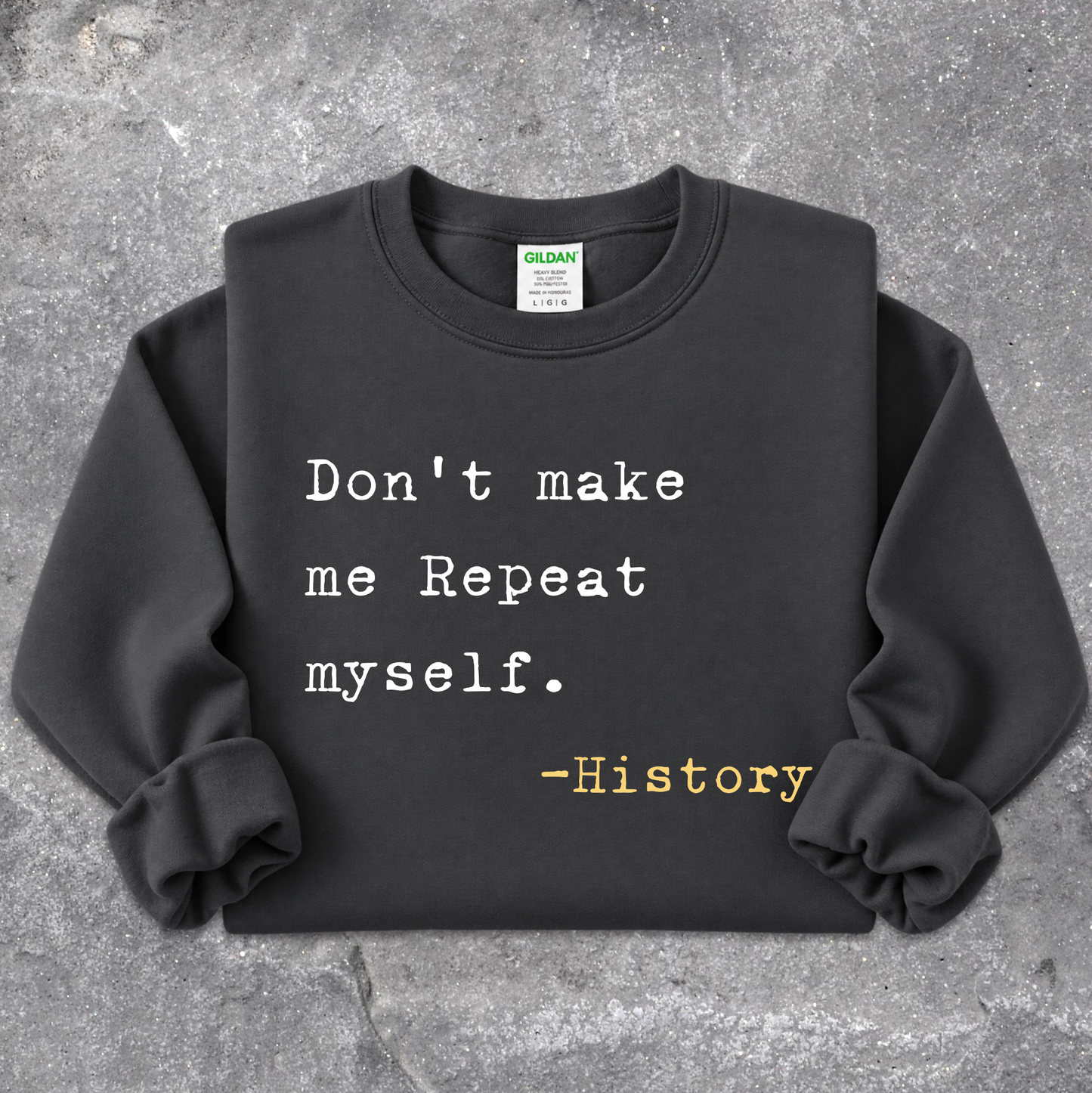 A black Gildan sweatshirt with rolled cuffs, featuring the printed text 'Don't make me Repeat myself. -History' in white and yellow on the front, laid flat on a textured gray concrete surface.