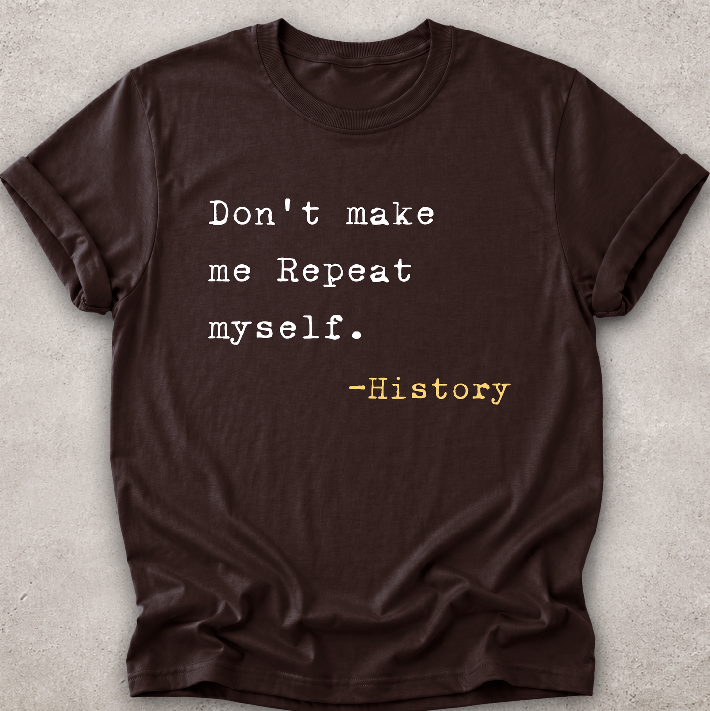 A dark brown short-sleeve t-shirt with a typewriter-style printed quote on the front reading 'Don't make me Repeat myself. -History' in white and yellow text, laid flat on a light gray textured background.