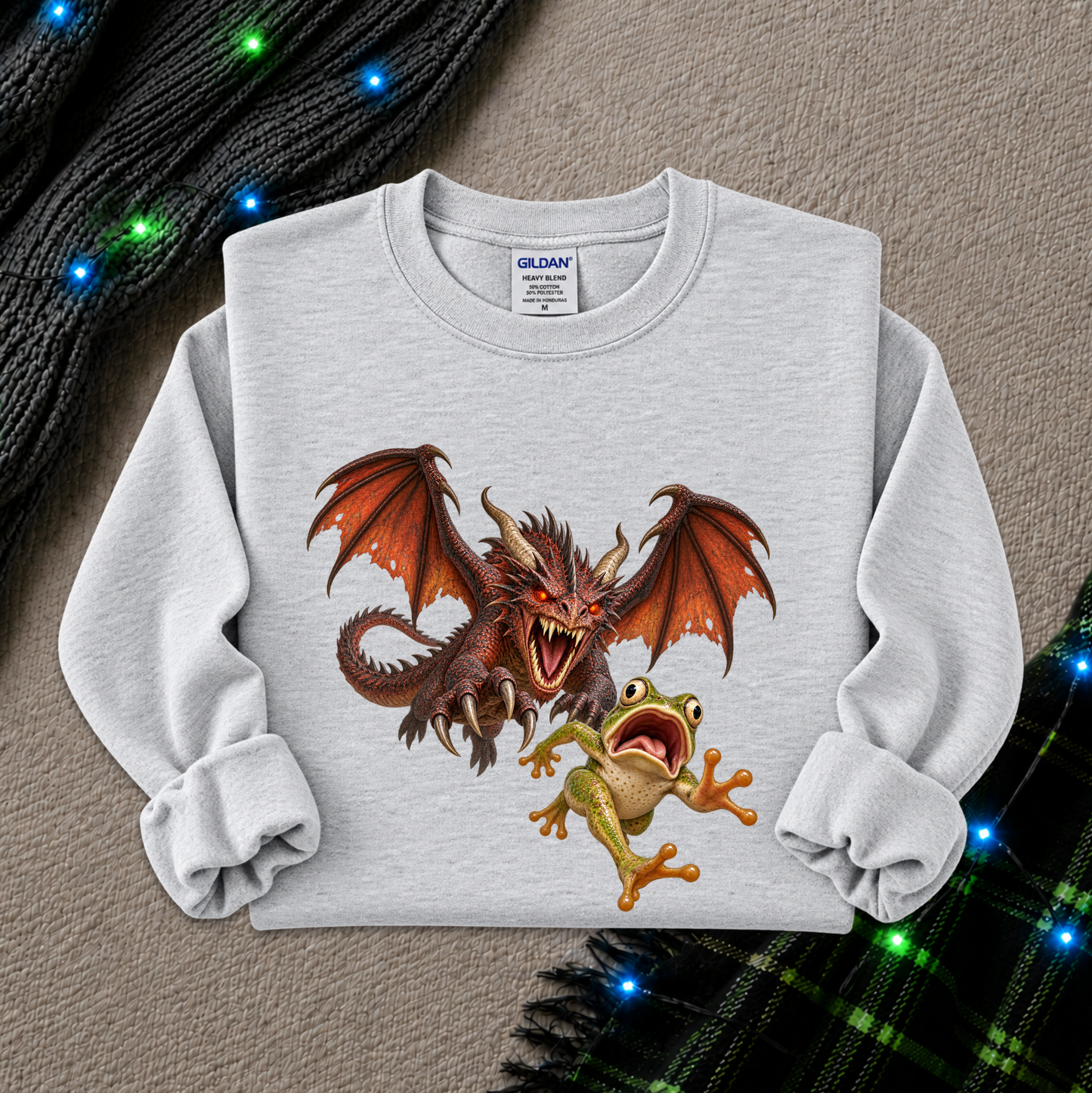 A light gray long-sleeve crewneck sweatshirt featuring a graphic print of a red dragon and a green frog, styled flat on a textured beige background with holiday lights and a plaid scarf.