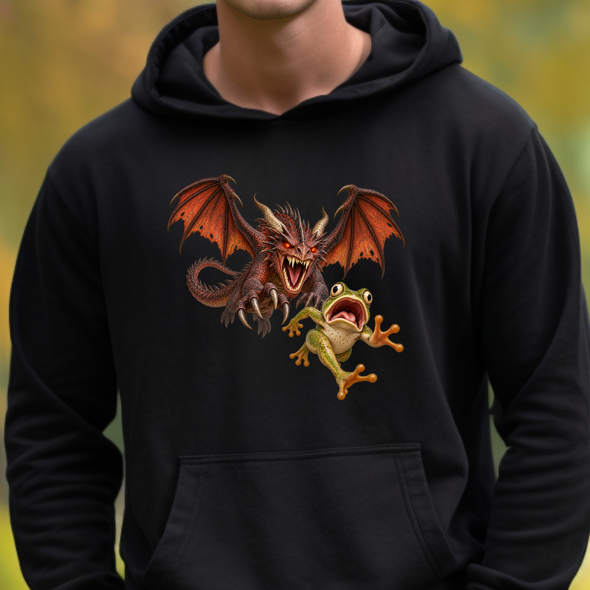 A person wearing a black pullover hoodie with a graphic print of a red dragon and a green tree frog facing off, set against a blurred yellow-green outdoor background.