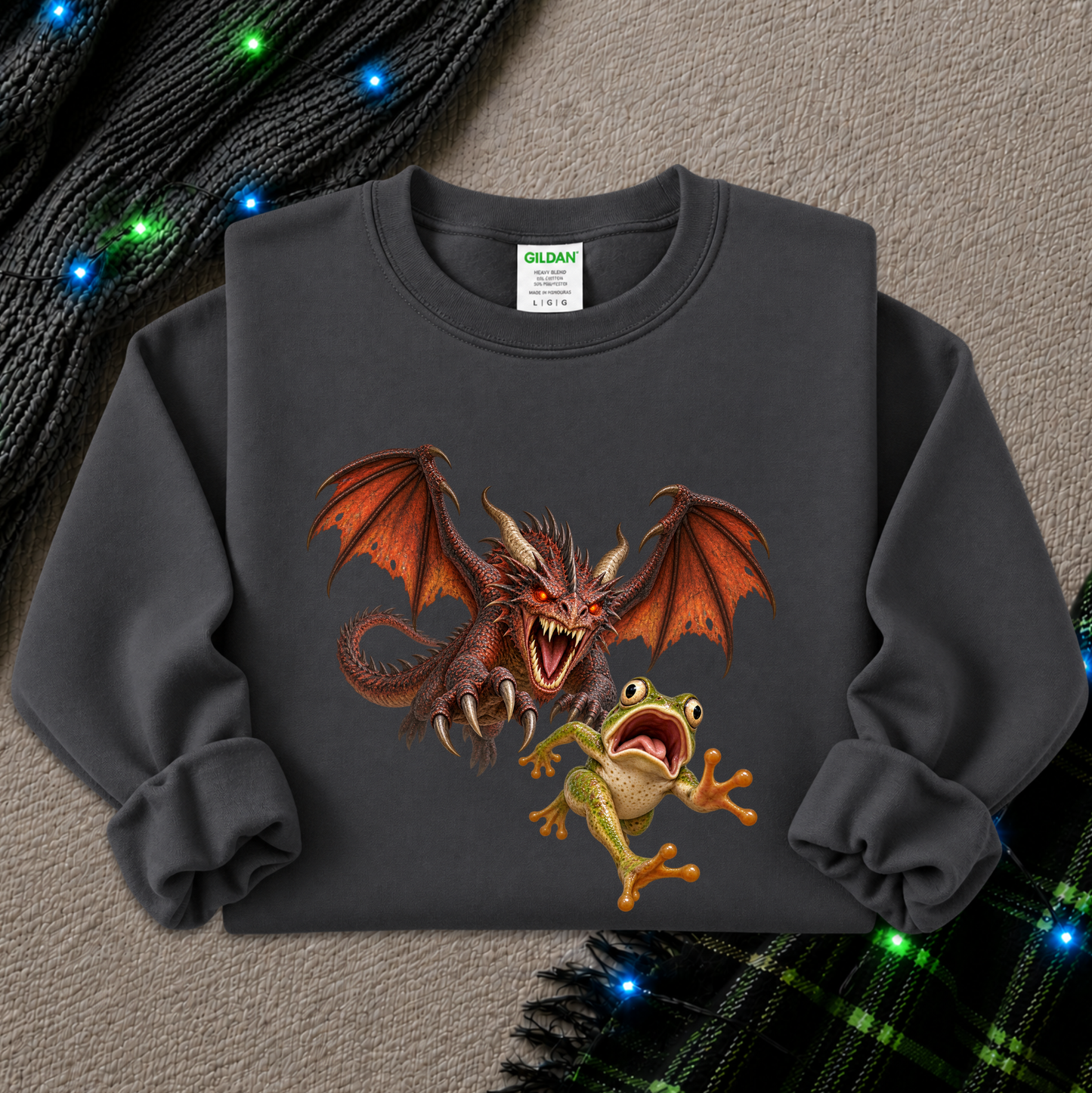 A dark gray crewneck sweatshirt featuring a graphic print of a red dragon and a green frog, styled flat on a textured gray background with holiday lights and plaid fabric accents.