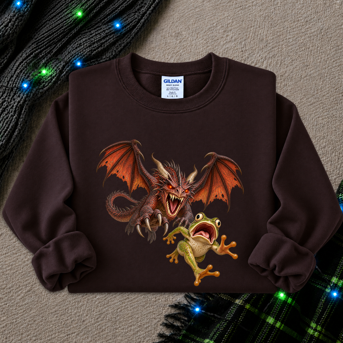 A dark brown crewneck sweatshirt featuring a graphic print of a red dragon and a green frog, styled flat on a textured beige background with holiday lights and plaid fabric accents.
