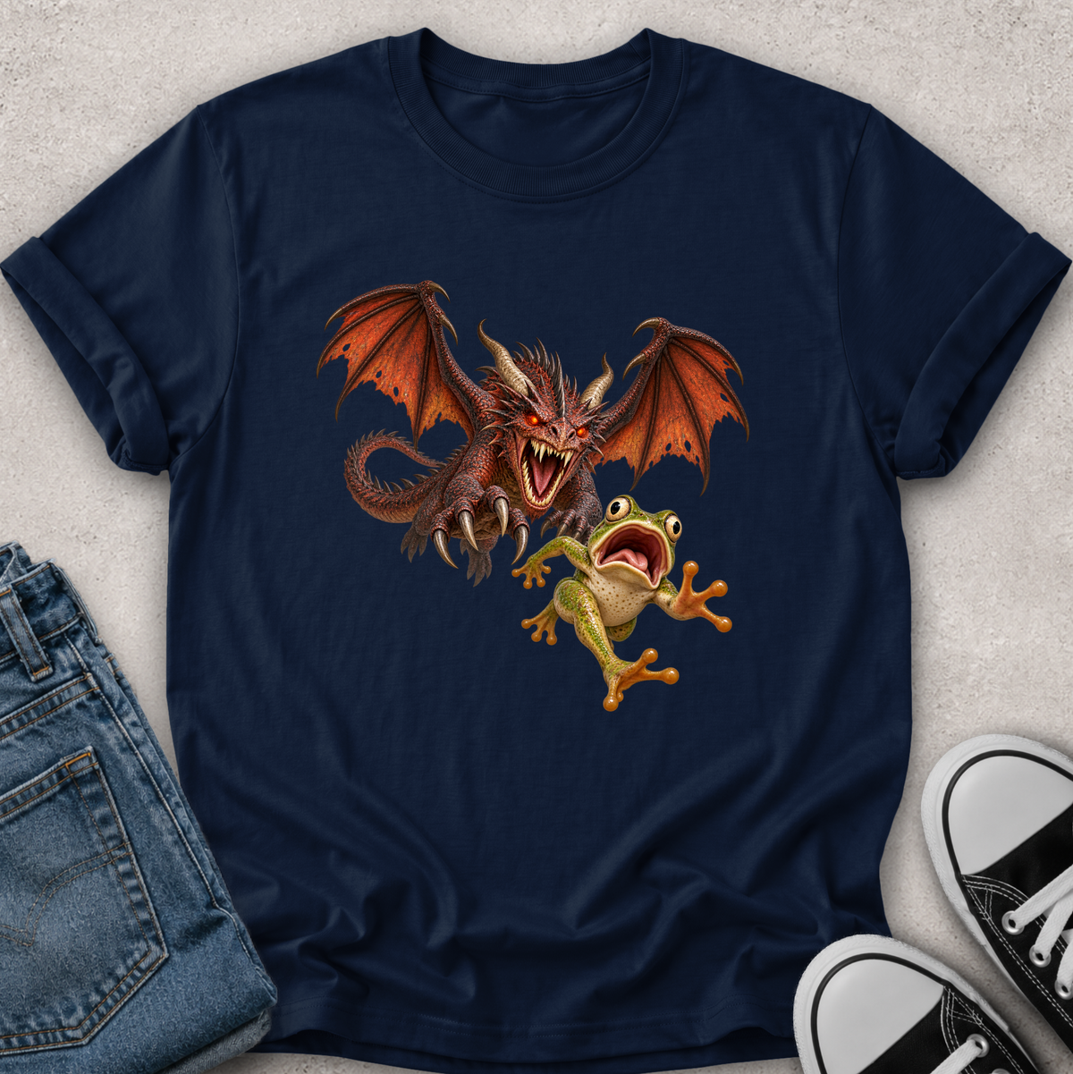 A navy blue short-sleeve t-shirt laid flat on a light gray textured surface, featuring a graphic print of a red dragon and a green tree frog. The shirt is styled with folded blue jeans on the left and black-and-white sneakers on the right.