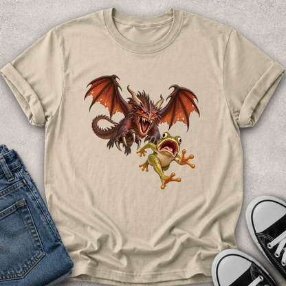 A beige graphic t-shirt laid flat on a light gray textured surface, styled with folded blue jeans and black-and-white sneakers. The t-shirt features a graphic print of a brown dragon with orange wings facing off against a green tree frog.