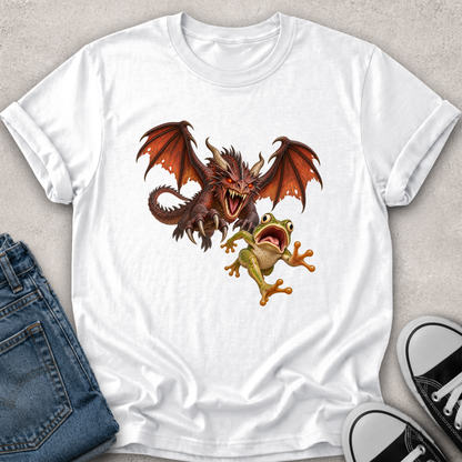 A white short-sleeve crewneck t-shirt laid flat on a light gray textured surface, featuring a graphic print of a red-brown dragon and a green tree frog. The shirt is styled with folded blue jeans on the left and black-and-white sneakers on the right.
