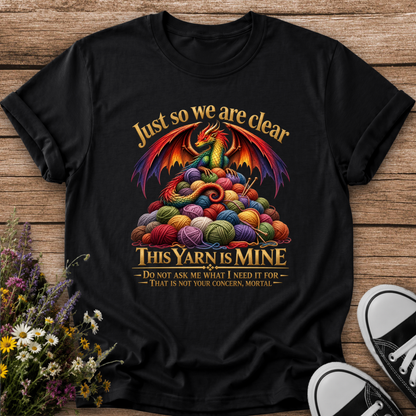 A black graphic t-shirt laid flat on a rustic wooden surface, styled with a small bouquet of wildflowers on the bottom left and black-and-white sneakers on the bottom right. The t-shirt features a colorful graphic of a dragon wrapped in knitting yarn with the text 'Just so we are clear THIS YARN IS MINE'.