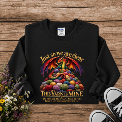 A black crewneck sweatshirt with a colorful graphic of a dragon rising from a pile of knitting yarn balls, with the text 'Just so we are clear THIS YARN IS MINE'. The sweatshirt is laid flat on a rustic wooden background with flowers and sneakers as props.
