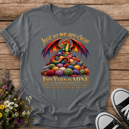 A dark gray graphic t-shirt laid flat on a rustic wooden surface, styled with wildflowers on the left and black-and-white sneakers on the right. The t-shirt features a colorful graphic of a red and green dragon wrapped in a pile of multicolored yarn, with the text 'Just so we are clear THIS YARN IS MINE'.