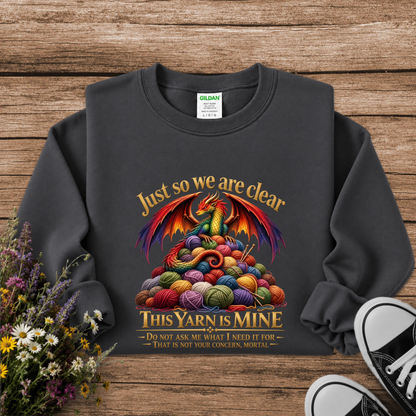 A dark charcoal gray crewneck sweatshirt with a colorful graphic of a dragon rising from a pile of yarn balls, with the text 'Just so we are clear THIS YARN IS MINE'. The sweatshirt is displayed flat on a rustic wooden background with decorative flowers and black-and-white sneakers.