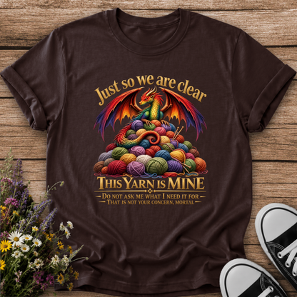 A dark brown graphic t-shirt laid flat on a rustic wooden background, styled with wildflowers on the left and black-and-white sneakers on the bottom right. The t-shirt features a colorful graphic of a dragon surrounded by balls of yarn with the text 'Just so we are clear THIS YARN IS MINE'.