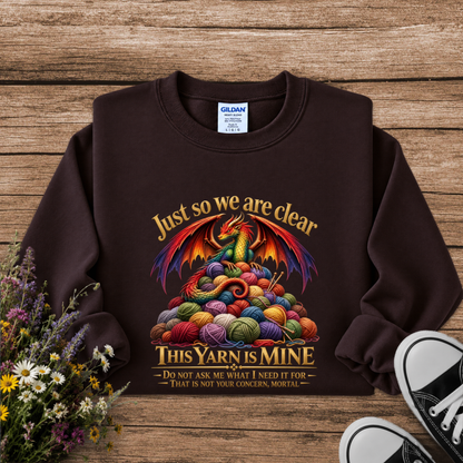 A dark brown crewneck sweatshirt with a colorful graphic of a dragon rising from a pile of knitting yarn balls, with the text 'Just so we are clear THIS YARN IS MINE'. The sweatshirt is displayed flat on a rustic wooden background with decorative flowers and black-and-white sneakers.