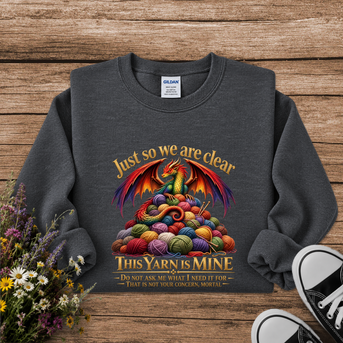 A dark gray crewneck sweatshirt laid flat on a rustic wooden surface, featuring a colorful graphic of a dragon rising from a pile of multicolored yarn balls with the text 'Just so we are clear THIS YARN IS MINE'. A Gildan brand tag is visible inside the collar.
