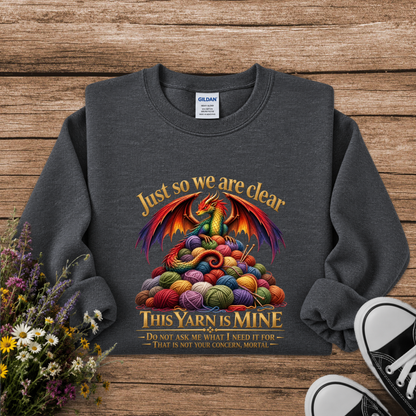 A dark gray crewneck sweatshirt laid flat on a rustic wooden surface, featuring a colorful graphic of a dragon rising from a pile of multicolored yarn balls with the text 'Just so we are clear THIS YARN IS MINE'. A Gildan brand tag is visible inside the collar.