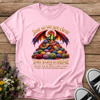 A light pink short-sleeve graphic t-shirt laid flat on a rustic wooden surface, styled with a small bouquet of wildflowers on the left and black-and-white sneakers on the right. The graphic features a colorful dragon rising from a pile of multicolored yarn balls with the text 'Just so we are clear THIS YARN IS MINE'.
