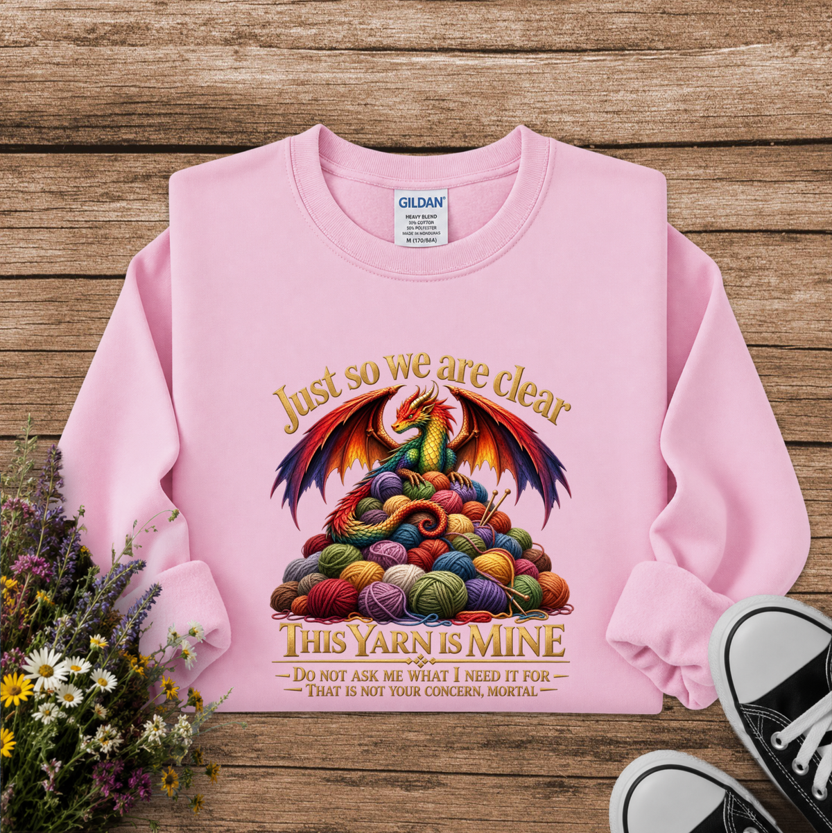A pink long-sleeve sweatshirt laid flat on a rustic wooden surface, featuring a colorful dragon made of yarn balls with the text 'Just so we are clear THIS YARN IS MINE.' A Gildan label is visible inside the collar, and flowers and black-and-white sneakers appear as styled props around the garment.