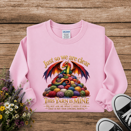 A pink long-sleeve sweatshirt laid flat on a rustic wooden surface, featuring a colorful dragon made of yarn balls with the text 'Just so we are clear THIS YARN IS MINE.' A Gildan label is visible inside the collar, and flowers and black-and-white sneakers appear as styled props around the garment.