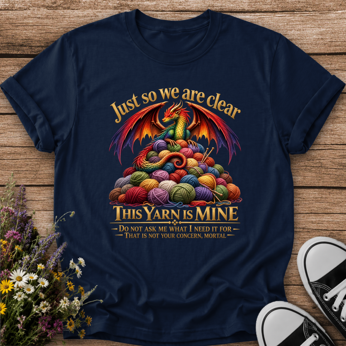 A navy blue graphic t-shirt laid flat on a rustic wooden surface, styled with a small bouquet of wildflowers on the left and black-and-white sneakers on the bottom right. The t-shirt features a colorful graphic of a green dragon wrapped in multicolored knitting yarn, with the text 'Just so we are clear THIS YARN IS MINE' and a smaller quote below.