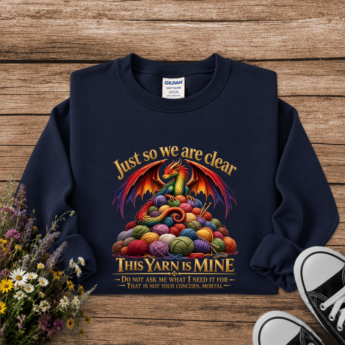 A navy blue crewneck sweatshirt with a colorful graphic of a dragon rising from a pile of knitting yarn balls, with the text 'Just so we are clear THIS YARN IS MINE'. The sweatshirt is laid flat on a rustic wooden background with wildflowers and black-and-white sneakers as props.
