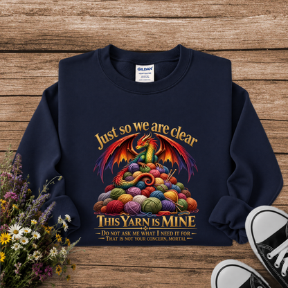 A navy blue crewneck sweatshirt with a colorful graphic of a dragon rising from a pile of knitting yarn balls, with the text 'Just so we are clear THIS YARN IS MINE'. The sweatshirt is laid flat on a rustic wooden background with wildflowers and black-and-white sneakers as props.