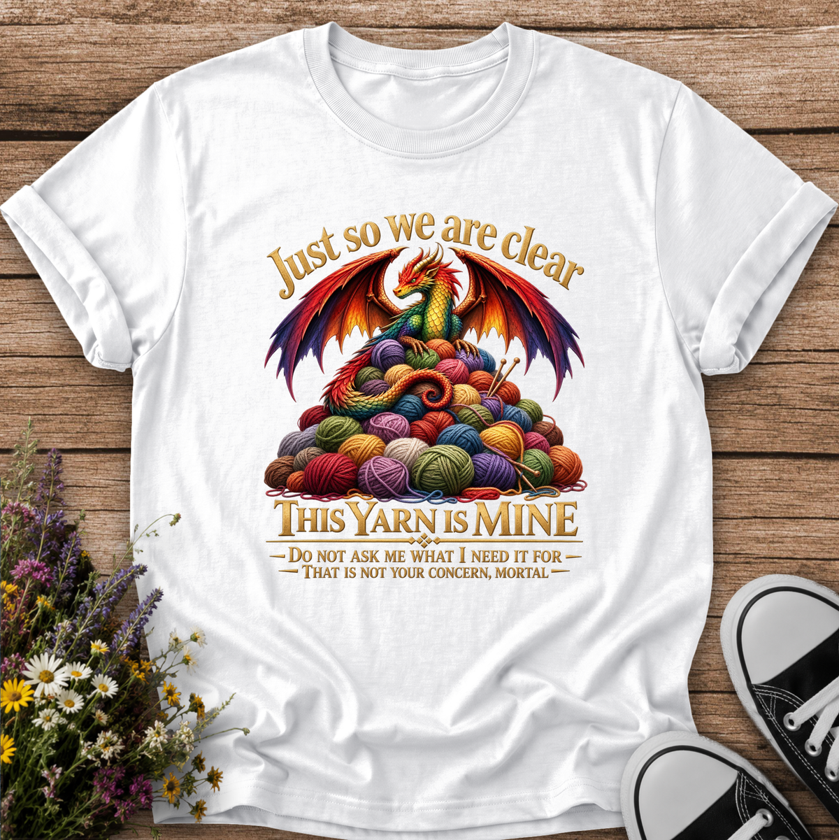 A white short-sleeve graphic t-shirt laid flat on a rustic wooden surface, styled with small flowers on the left and black-and-white sneakers at the bottom right. The shirt features a colorful illustration of a dragon rising from a pile of multicolored yarn balls, with the text 'Just so we are clear THIS YARN IS MINE' and a smaller warning below.