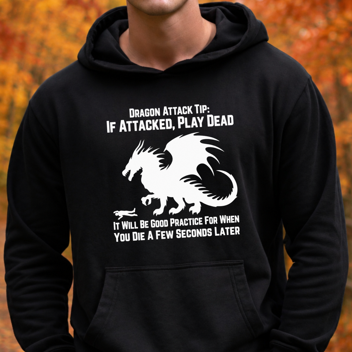 A person wearing a black pullover hoodie with a white graphic print of a dragon and humorous text about dragon attacks. The background shows blurred autumn foliage in orange and brown tones.