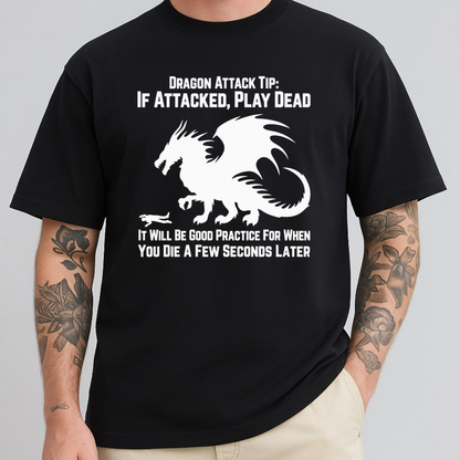 A man wearing a black short-sleeve graphic T-shirt with a white dragon illustration and text that reads, 'Dragon Attack Tip: IF ATTACKED, PLAY DEAD' and 'IT WILL BE GOOD PRACTICE FOR WHEN YOU DIE A FEW SECONDS LATER.' The photo is cropped from neck to upper thighs on a plain light gray studio background.