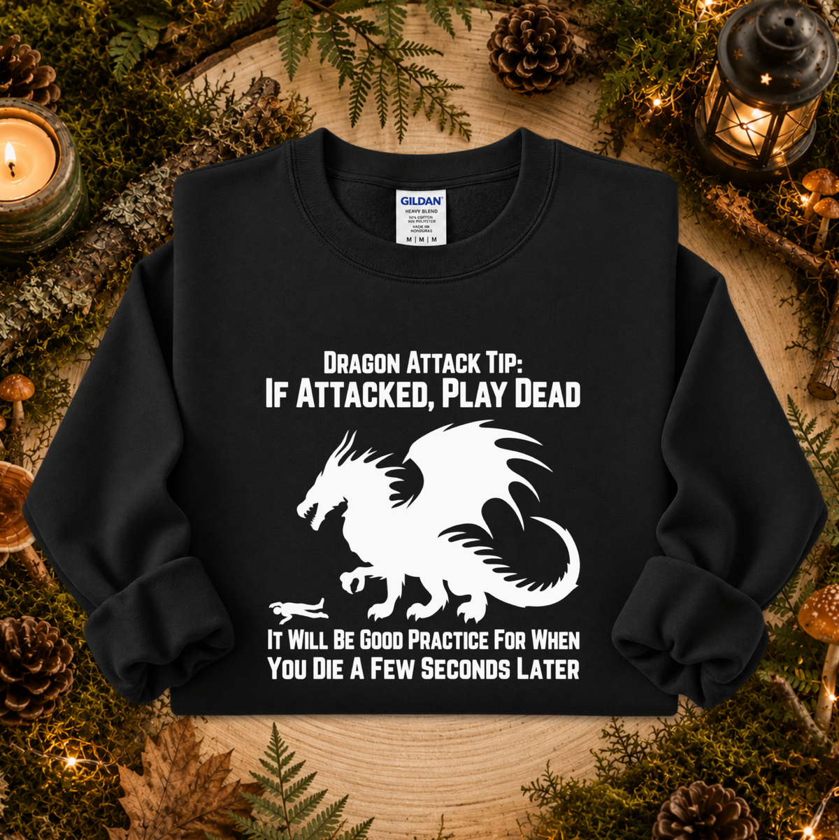 A black long-sleeve crewneck sweatshirt with a white graphic of a dragon and the text 'DRAGON ATTACK TIP: IF ATTACKED, PLAY DEAD' and 'IT WILL BE GOOD PRACTICE FOR WHEN YOU DIE A FEW SECONDS LATER'. The sweatshirt is displayed flat on a rustic wood background surrounded by pinecones, moss, and a candle.