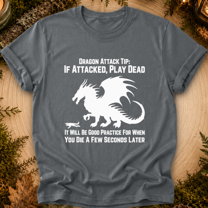 A dark gray graphic t-shirt laid flat on a wooden surface with mossy props. The shirt features a white dragon graphic and the text 'DRAGON ATTACK TIP: IF ATTACKED, PLAY DEAD' and 'IT WILL BE GOOD PRACTICE FOR WHEN YOU DIE A FEW SECONDS LATER'.