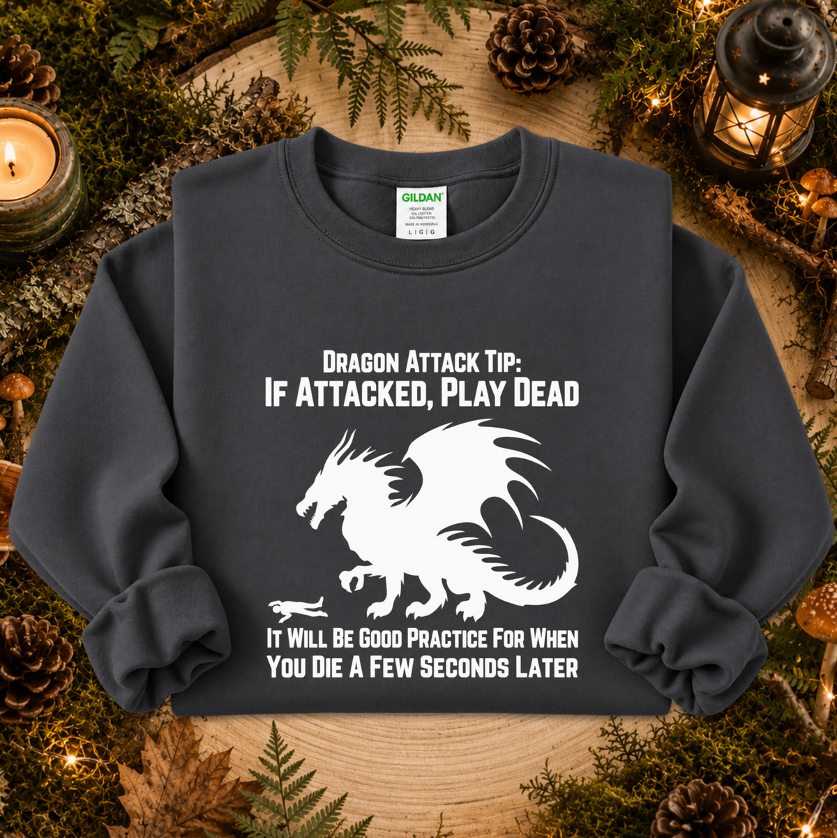 A dark charcoal gray crewneck sweatshirt with a white graphic of a dragon and humorous text, displayed in a cozy forest-themed flat lay with pinecones, moss, and a candle.