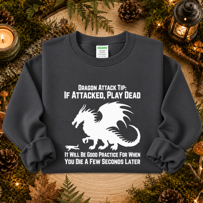 A dark charcoal gray crewneck sweatshirt with a white graphic of a dragon and humorous text, displayed in a cozy forest-themed flat lay with pinecones, moss, and a candle.