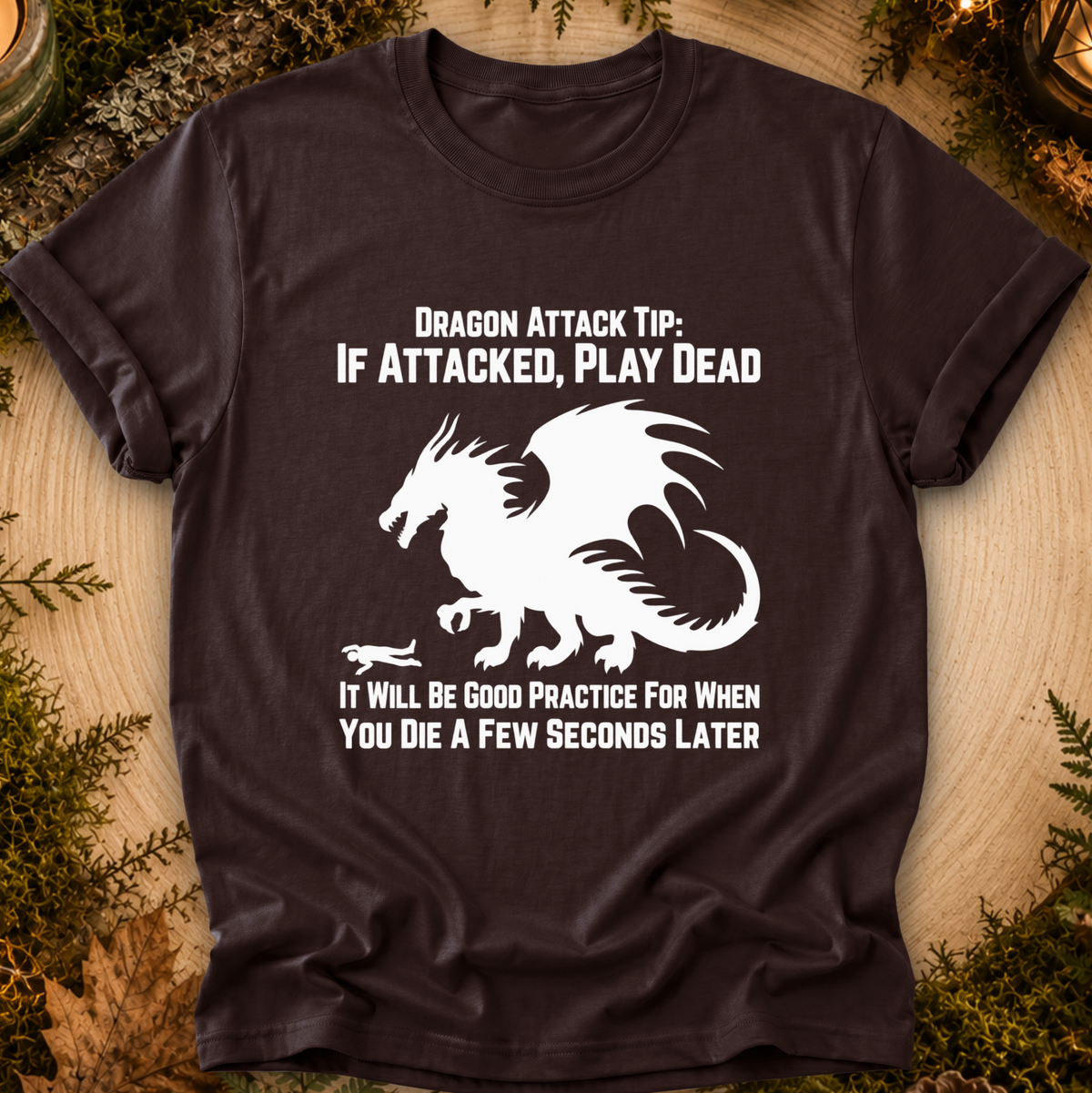 A dark brown short-sleeve t-shirt with a white graphic of a dragon and the text 'DRAGON ATTACK TIP: IF ATTACKED, PLAY DEAD' and 'IT WILL BE GOOD PRACTICE FOR WHEN YOU DIE A FEW SECONDS LATER', laid flat on a wood surface with decorative greenery around the edges.
