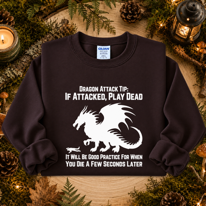 A dark brown crewneck sweatshirt featuring a white graphic of a dragon and the text 'DRAGON ATTACK TIP: IF ATTACKED, PLAY DEAD' and 'IT WILL BE GOOD PRACTICE FOR WHEN YOU DIE A FEW SECONDS LATER'. The sweatshirt is displayed flat on a rustic wooden background decorated with pinecones, moss, and a lit candle.