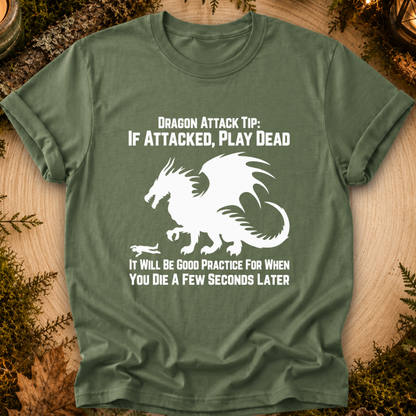 An olive green short-sleeve t-shirt with a white graphic of a dragon and humorous text, laid flat on a rustic wood background with mossy accents.