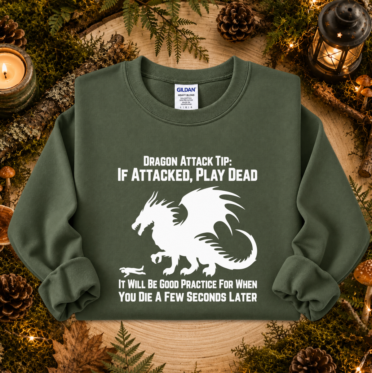 An olive green crewneck sweatshirt with a white graphic of a dragon and humorous text, displayed in a cozy flat-lay setting with pinecones, moss, and candles.