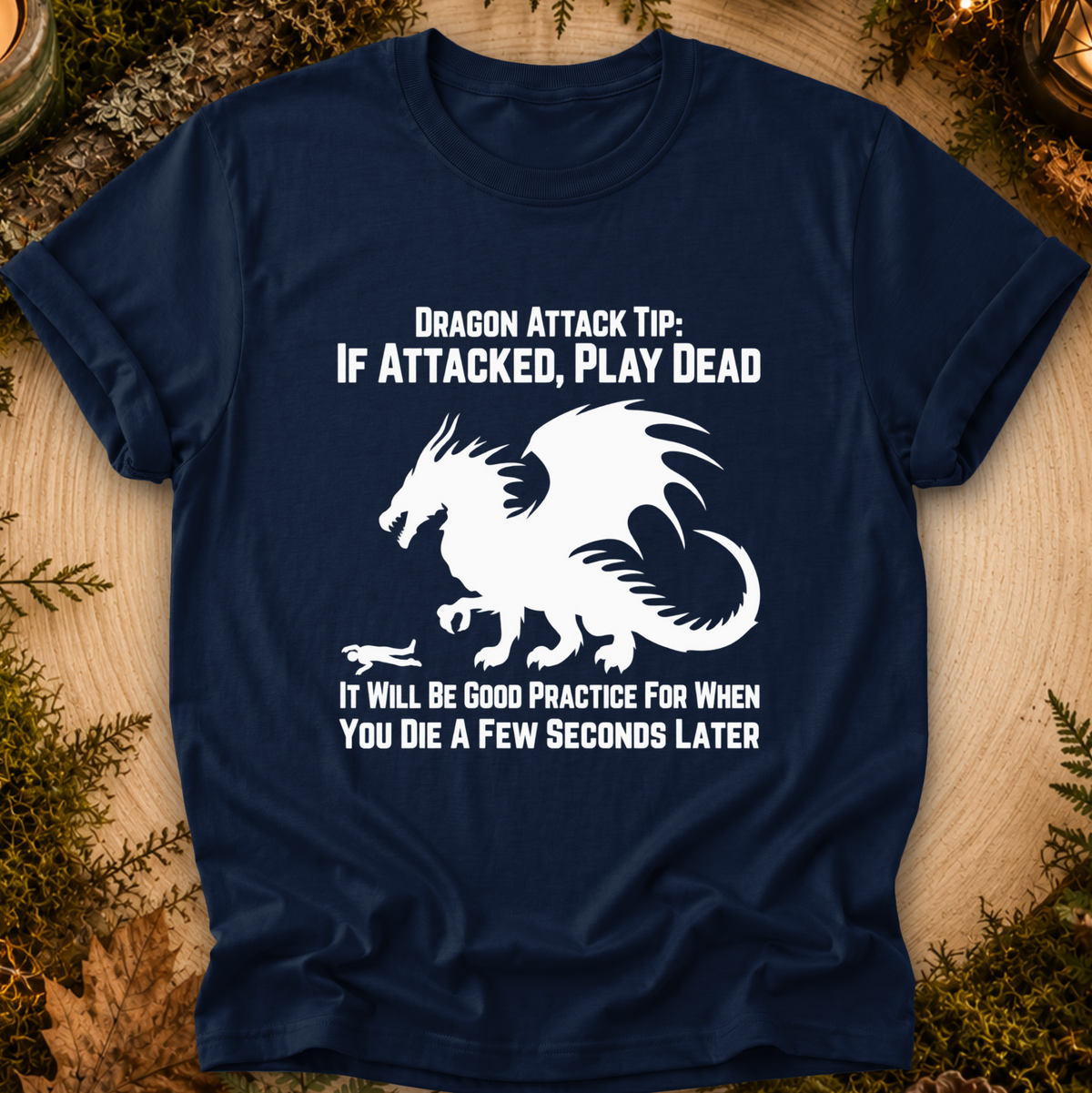 A navy blue graphic t-shirt laid flat on a rustic wood background with greenery. The shirt features a white dragon illustration and the text 'DRAGON ATTACK TIP: IF ATTACKED, PLAY DEAD' and 'IT WILL BE GOOD PRACTICE FOR WHEN YOU DIE A FEW SECONDS LATER'.