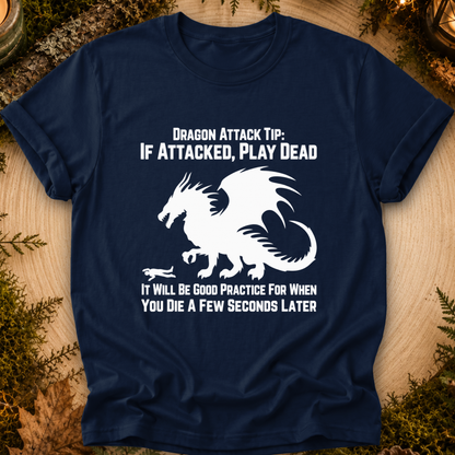 A navy blue graphic t-shirt laid flat on a rustic wood background with greenery. The shirt features a white dragon illustration and the text 'DRAGON ATTACK TIP: IF ATTACKED, PLAY DEAD' and 'IT WILL BE GOOD PRACTICE FOR WHEN YOU DIE A FEW SECONDS LATER'.