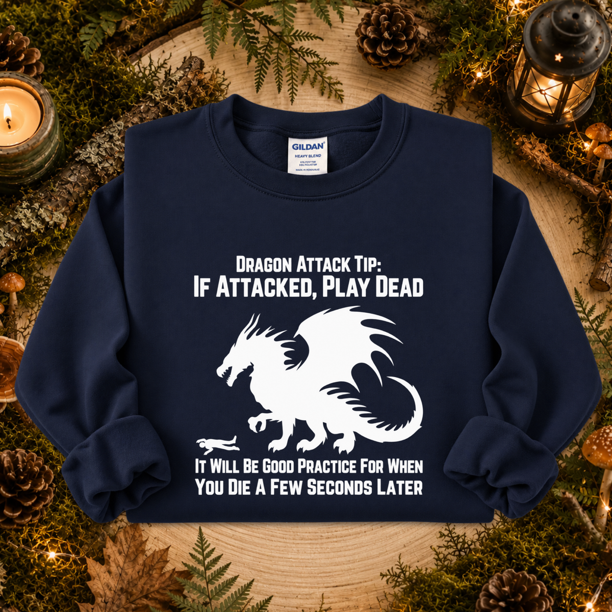 A navy blue crewneck sweatshirt with a white graphic of a dragon and the text 'DRAGON ATTACK TIP: IF ATTACKED, PLAY DEAD' and 'IT WILL BE GOOD PRACTICE FOR WHEN YOU DIE A FEW SECONDS LATER'. The sweatshirt is displayed flat on a rustic wooden surface surrounded by pinecones, moss, and candles.