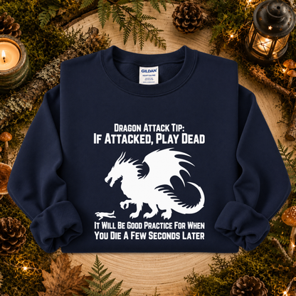 A navy blue crewneck sweatshirt with a white graphic of a dragon and the text 'DRAGON ATTACK TIP: IF ATTACKED, PLAY DEAD' and 'IT WILL BE GOOD PRACTICE FOR WHEN YOU DIE A FEW SECONDS LATER'. The sweatshirt is displayed flat on a rustic wooden surface surrounded by pinecones, moss, and candles.
