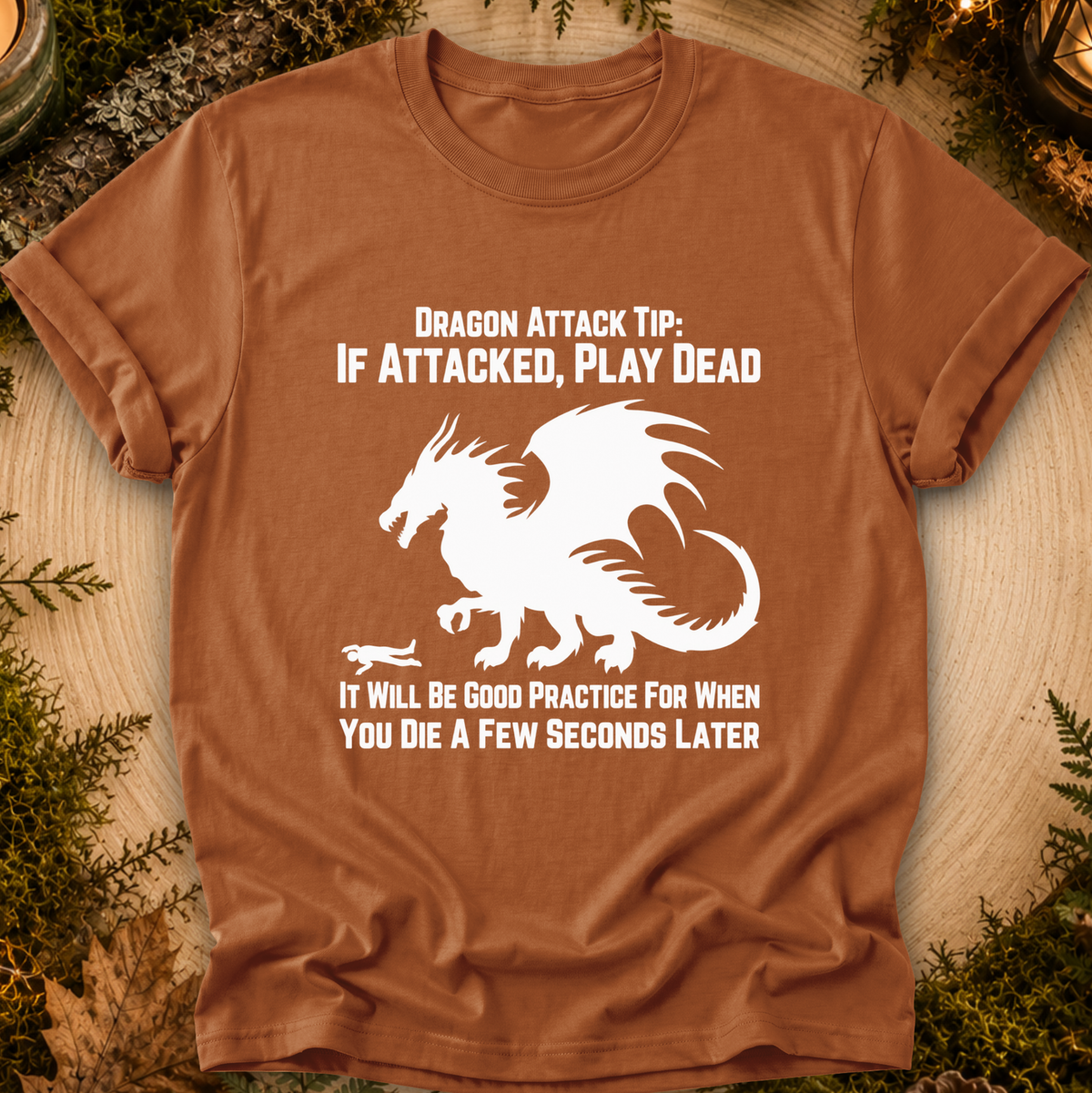 A brown short-sleeve graphic T-shirt laid flat on a wood surface with greenery around the edges. The shirt has a large white dragon illustration and the text: 'DRAGON ATTACK TIP: IF ATTACKED, PLAY DEAD' and 'IT WILL BE GOOD PRACTICE FOR WHEN YOU DIE A FEW SECONDS LATER'.