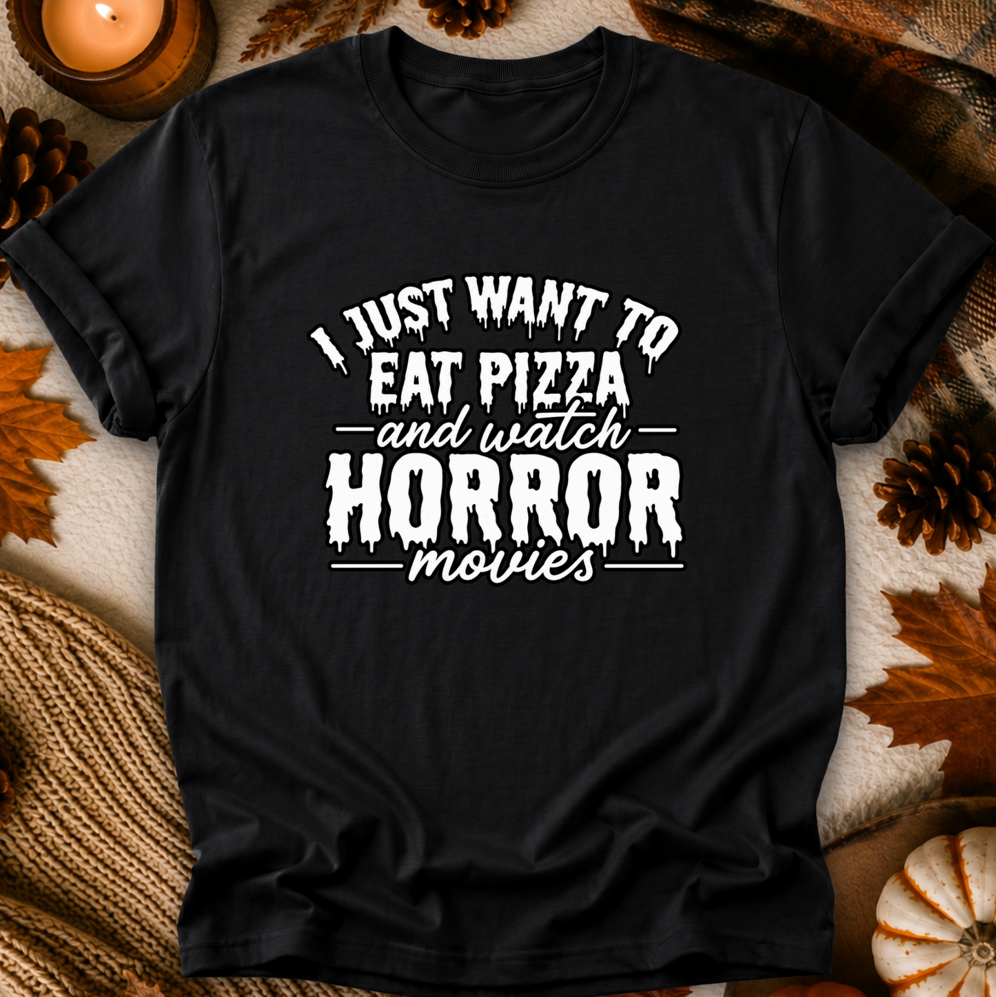 A black short-sleeve graphic T-shirt laid flat with a white horror-themed slogan reading 'I JUST WANT TO EAT PIZZA and watch HORROR movies.' The shirt is styled in a cozy autumn flat lay with leaves, pinecones, a candle, and a small pumpkin around it.