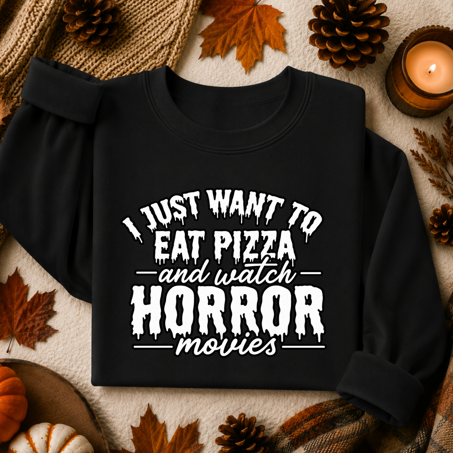 A black long-sleeve sweatshirt with rolled cuffs and a white graphic text print reading 'I JUST WANT TO EAT PIZZA and watch HORROR movies', styled in a flat lay with autumn leaves, pinecones, a candle, and a knit blanket.
