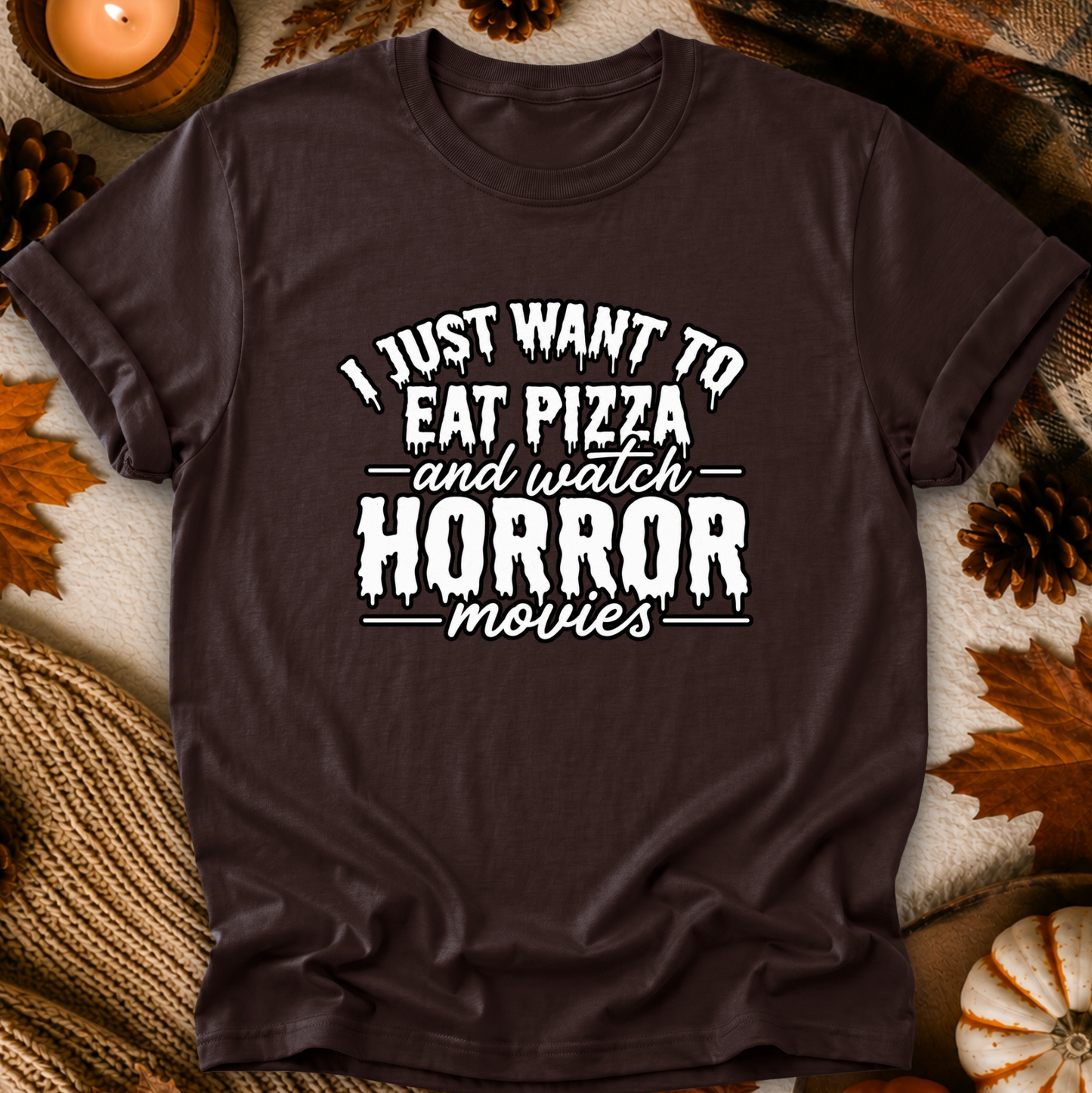 A dark brown short-sleeve t-shirt with a white graphic text reading 'I JUST WANT TO EAT PIZZA and watch HORROR movies' laid flat on a cozy autumn-themed background with leaves, pinecones, a candle, and a knit blanket.