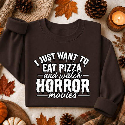 A dark brown sweatshirt with rolled-up sleeves, featuring a white graphic text that reads 'I JUST WANT TO EAT PIZZA and watch HORROR movies'. The sweatshirt is presented in a flat-lay arrangement with autumn props including pinecones, maple leaves, a candle, and a knit sweater.