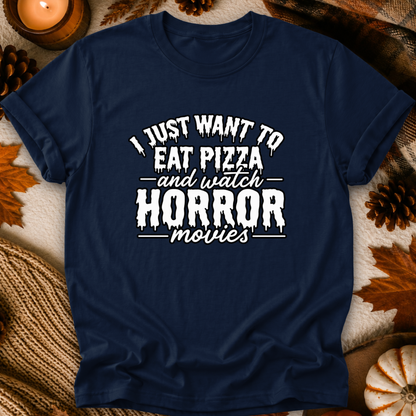A navy blue short-sleeve t-shirt with a white graphic text print reading 'I JUST WANT TO EAT PIZZA and watch HORROR movies', displayed in a flat-lay autumn setting with pinecones, leaves, and a candle.
